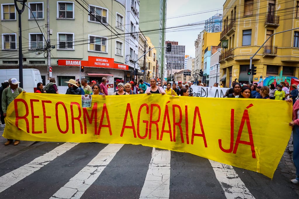 pessoas segurando cartaz amarelo na rua pedindo reforça agrária