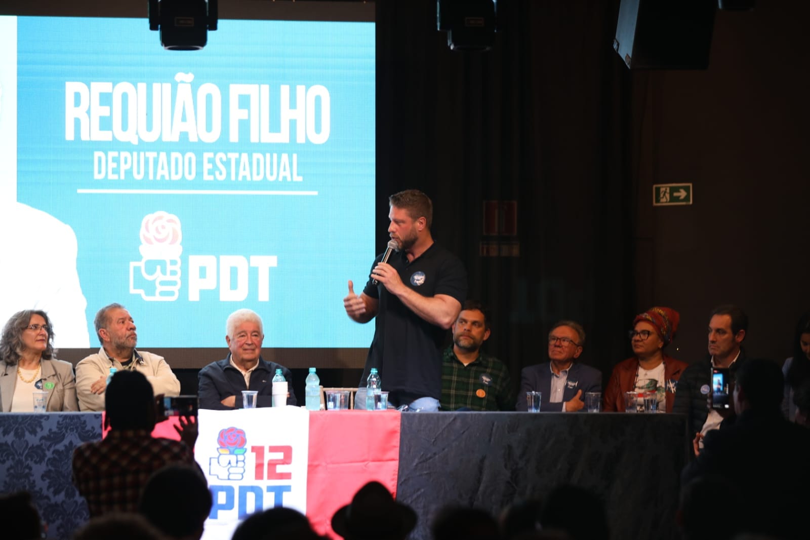 o deputado requião filho, homem branco de camisa polo preta segurando microfone e outras pessoas sentadas ao lado