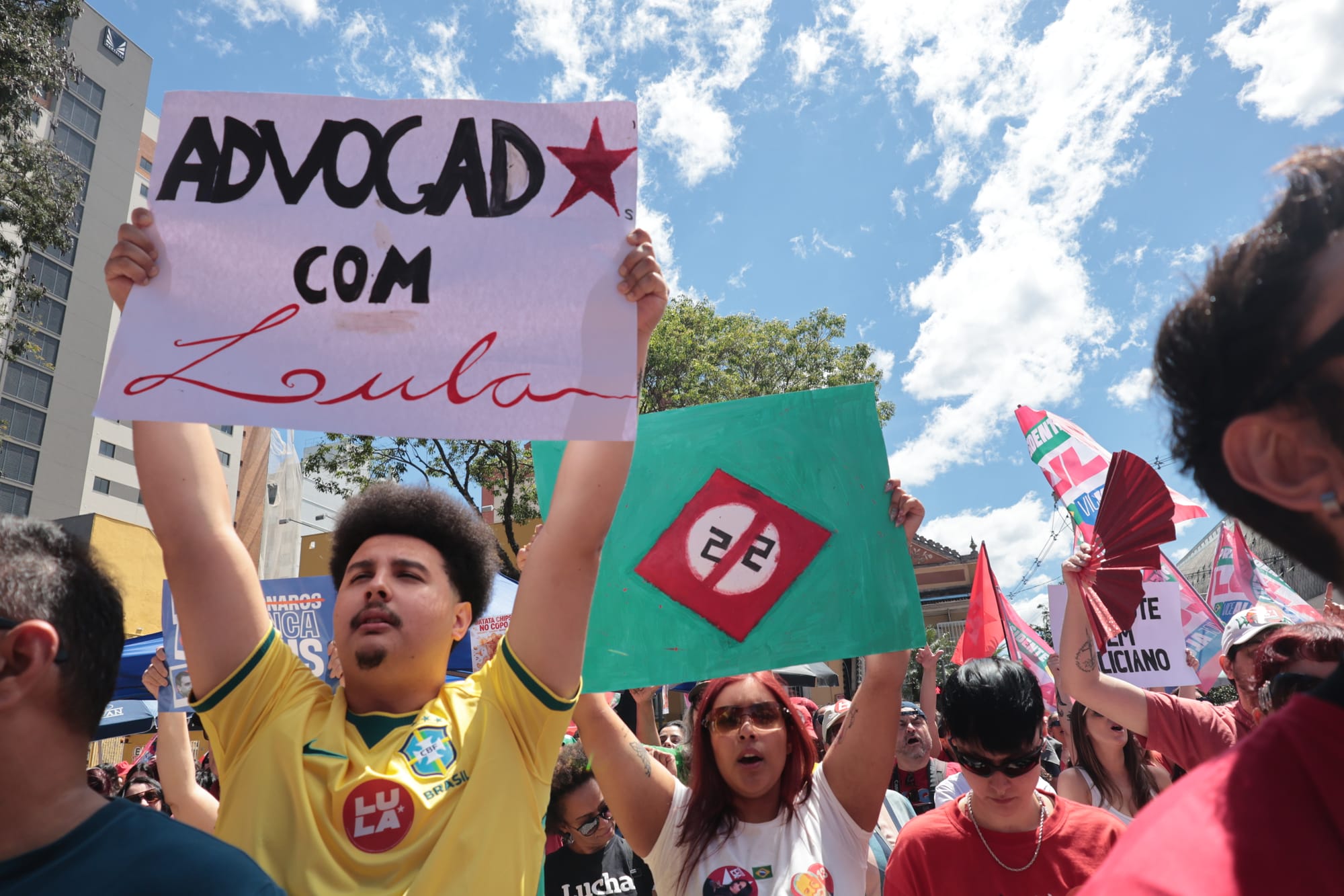 manifestantes segurança cartazes: mulher com cartal com número vinte e dois bloqueado e homem com cartaz escrito advogado com Lula