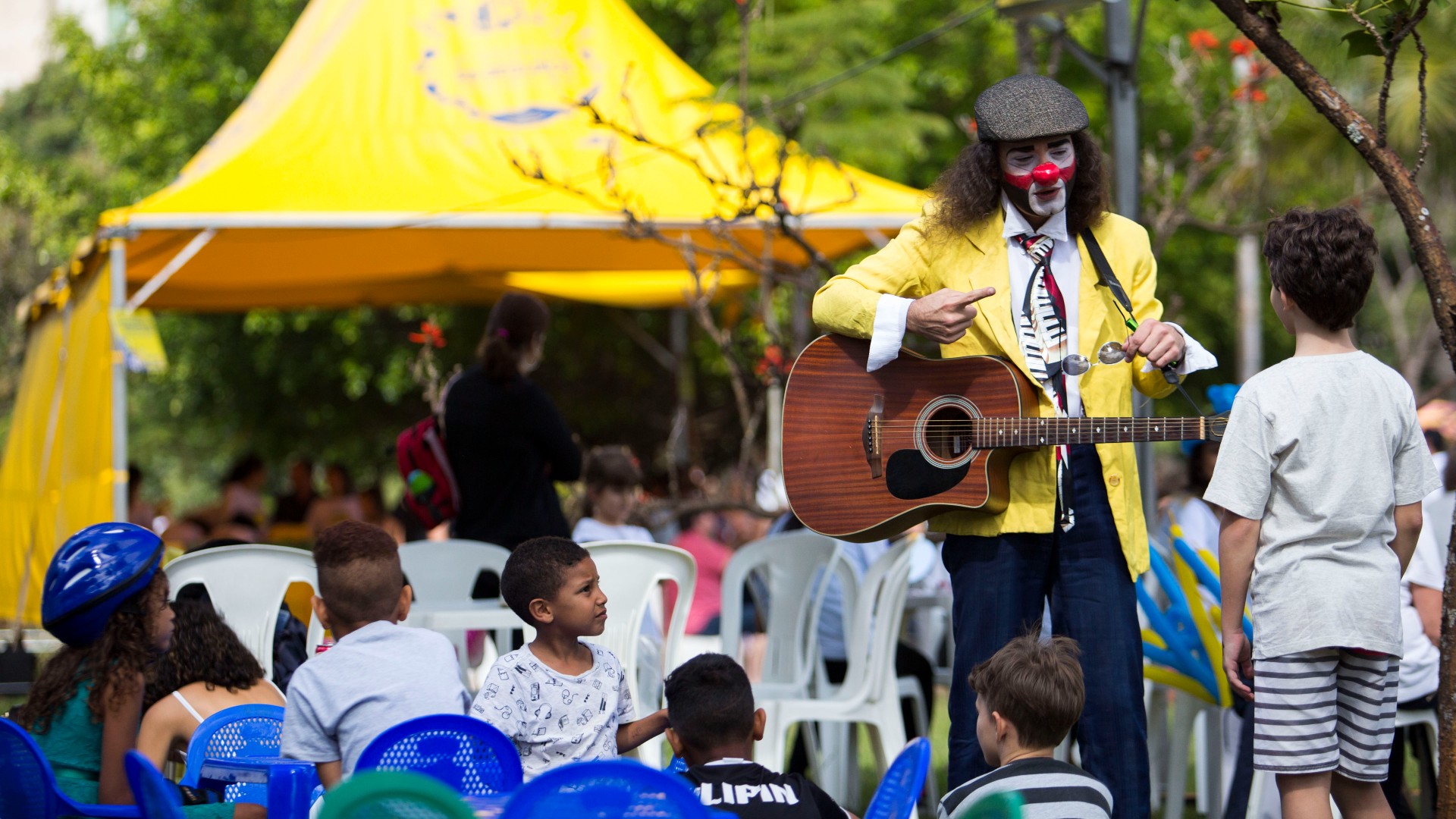#paratodosverem Imagem mostra um palhaço com um violão realizando atividade com crianças no Blue Jazz Brasil Festival.